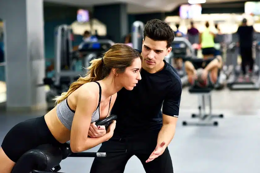 Personal trainer helping young woman lift weights while working out in a gym Top 4 Benefits Of Hiring A Personal Trainer